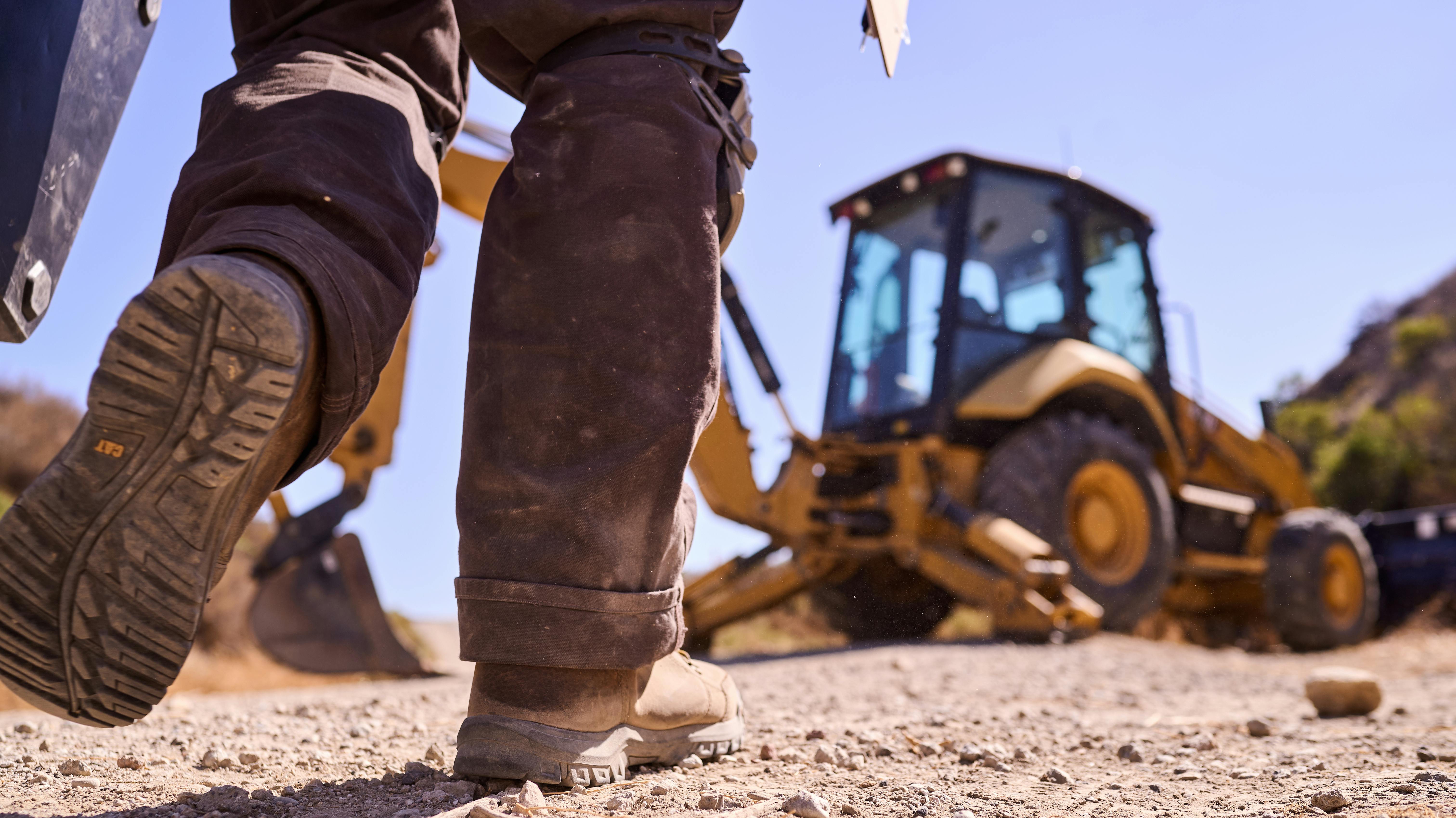 Man walking toward a Caterpillar backhoe in the dirt.