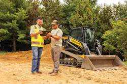 Two people looking at myNewHollandConstruction telematics platform on a screen. A New Holland compact track loader sits in the background.