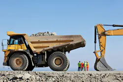 A giant dump truck sits near an excavator while men talk.