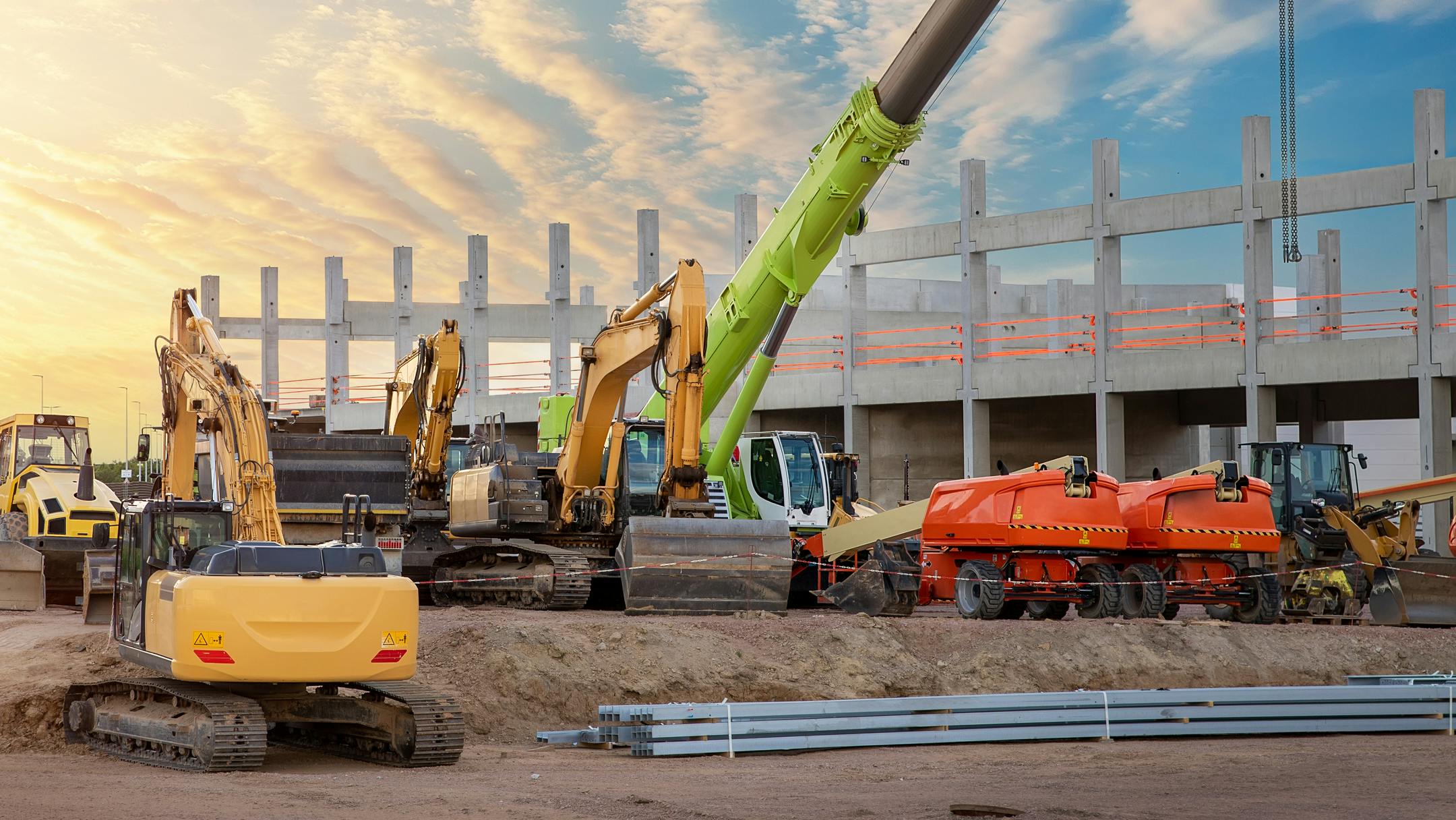 Many different multiclored colorful heavy industrial machinery equipment at construction site parking area against warehouse building city infrastructure development. Commercial vehicles rental sale.