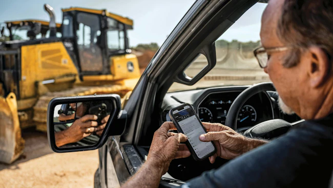 John Deere Operations Center. Man checks his phone while looking at a John Deere dozer.