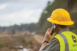 Contractor with a hardhat is on the phone. He's wearing a John Deere safety vest.