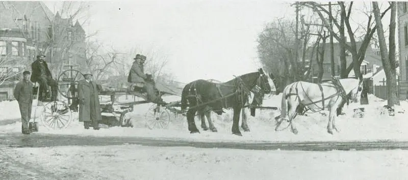 An old tyme photo of a horse drawn grader moving snow.