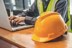 A yellow safety helmet sits next to a man in the construction industry, working on a computer..