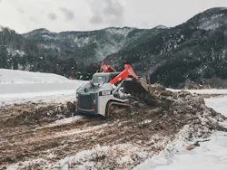 A Takeuchi compact track loader digs dirt in snowy mountains.